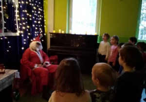 Mikołaj siedzi na fotelu. Dzieci stoją przed nim i śpiewają piosenkę